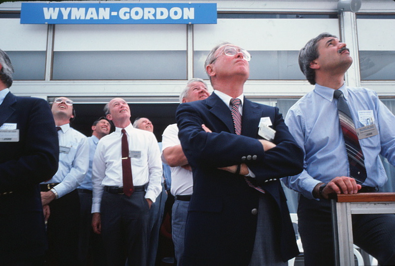 Two men with a crowd behind them, 
	looking up at planes flying in the air show demonstration.